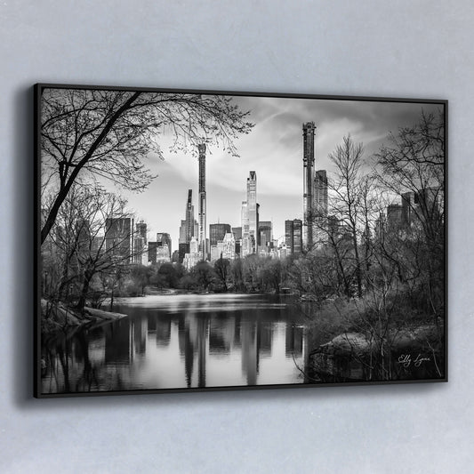 Black and white photograph of New York City skyline reflected in Central Park lake, framed by bare winter trees.